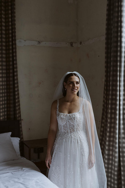 Bride stands next to bed in Lotter Dress and Pippa Veil with beaded border.