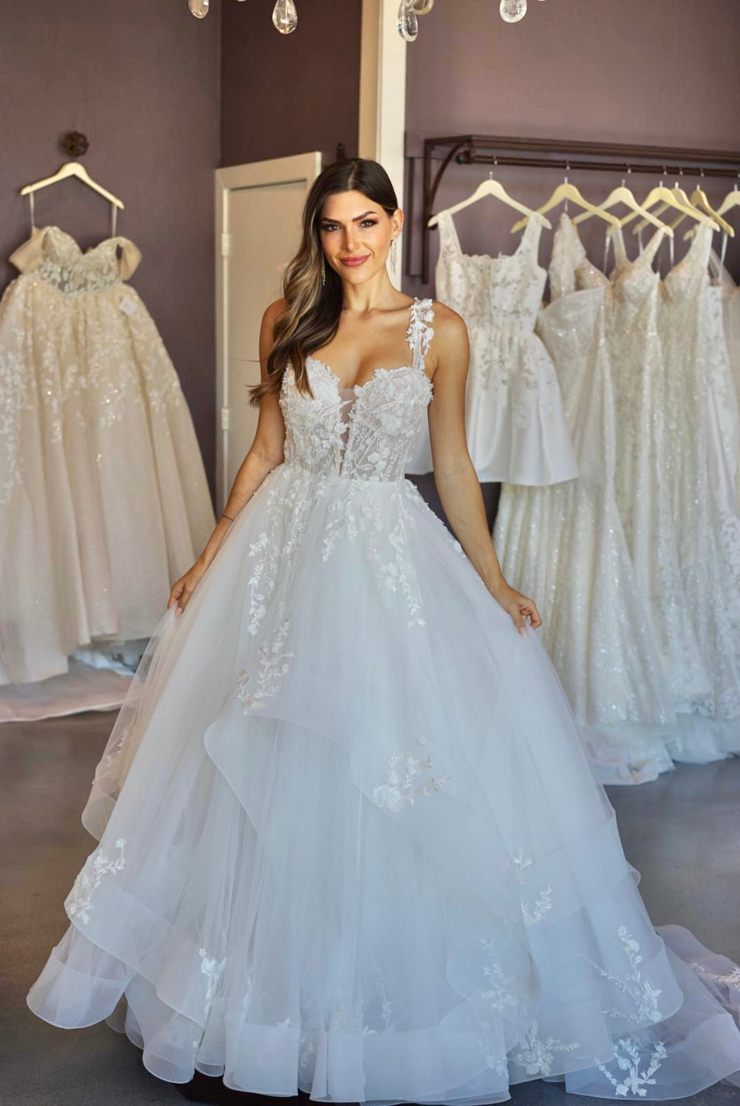 Tess is an a-line wedding gown with plunging sweetheart neckline. Asymmetrical tulle skirt creates volume. Delicate floral off-the-shoulder straps match the lace bodice and applique on skirt.