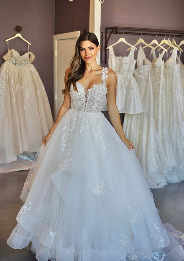 Tess is an a-line wedding gown with plunging sweetheart neckline. Asymmetrical tulle skirt creates volume. Delicate floral off-the-shoulder straps match the lace bodice and applique on skirt.