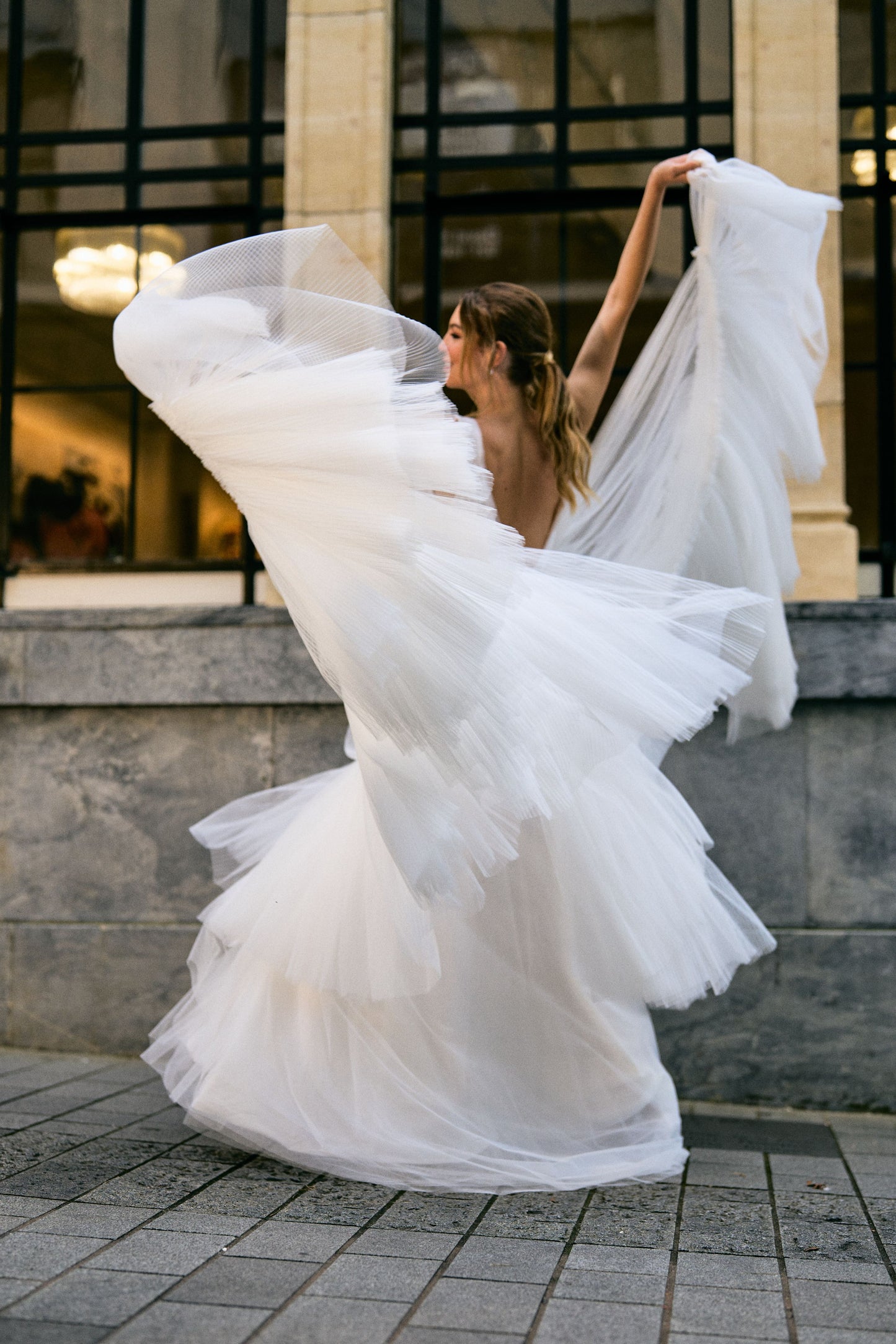 Rhia wedding dress with sheer pleated French Tulle fabric and A-line silhouette. Plunging v-neckline with floral lace overlay for a romantic touch. Two-tiered skirt adds volume and movement. Feminine and modern gown that gives the illusion of floating when walking down the aisle.