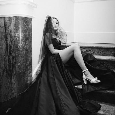 Model sits on marble stairs wearing black Daisha wedding dress with dramatic split.