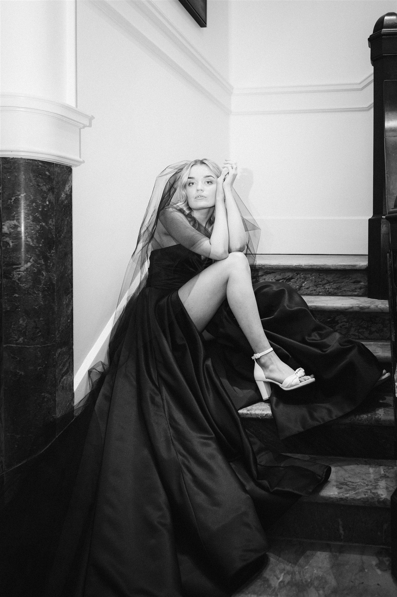 Model wears black satin gown and black veil. She sits on marble stairs.