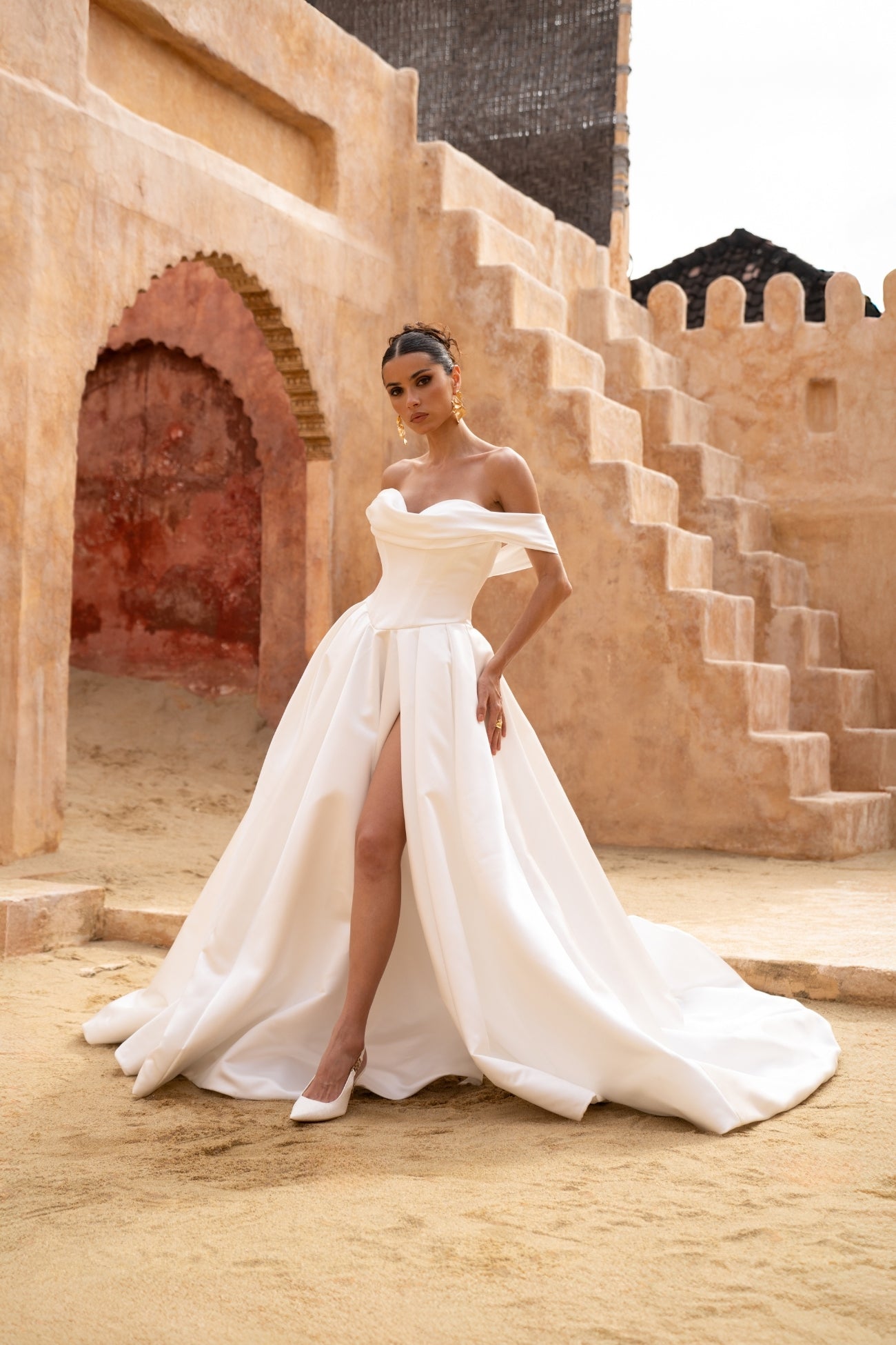 A line wedding gown featuring off the shoulder straps and a sweetheart neckline, in a minmal plain fabric, high split and dramatic skirt. 