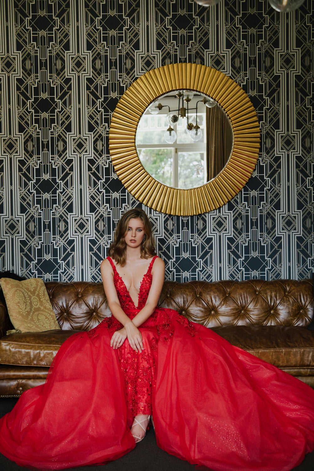Model wears red Nisha gown and overskirt sitting on a couch with layers of tulle splayed out beside her.