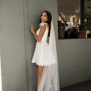 Bride stands against wall in white mini dress with long 3D floral veil.