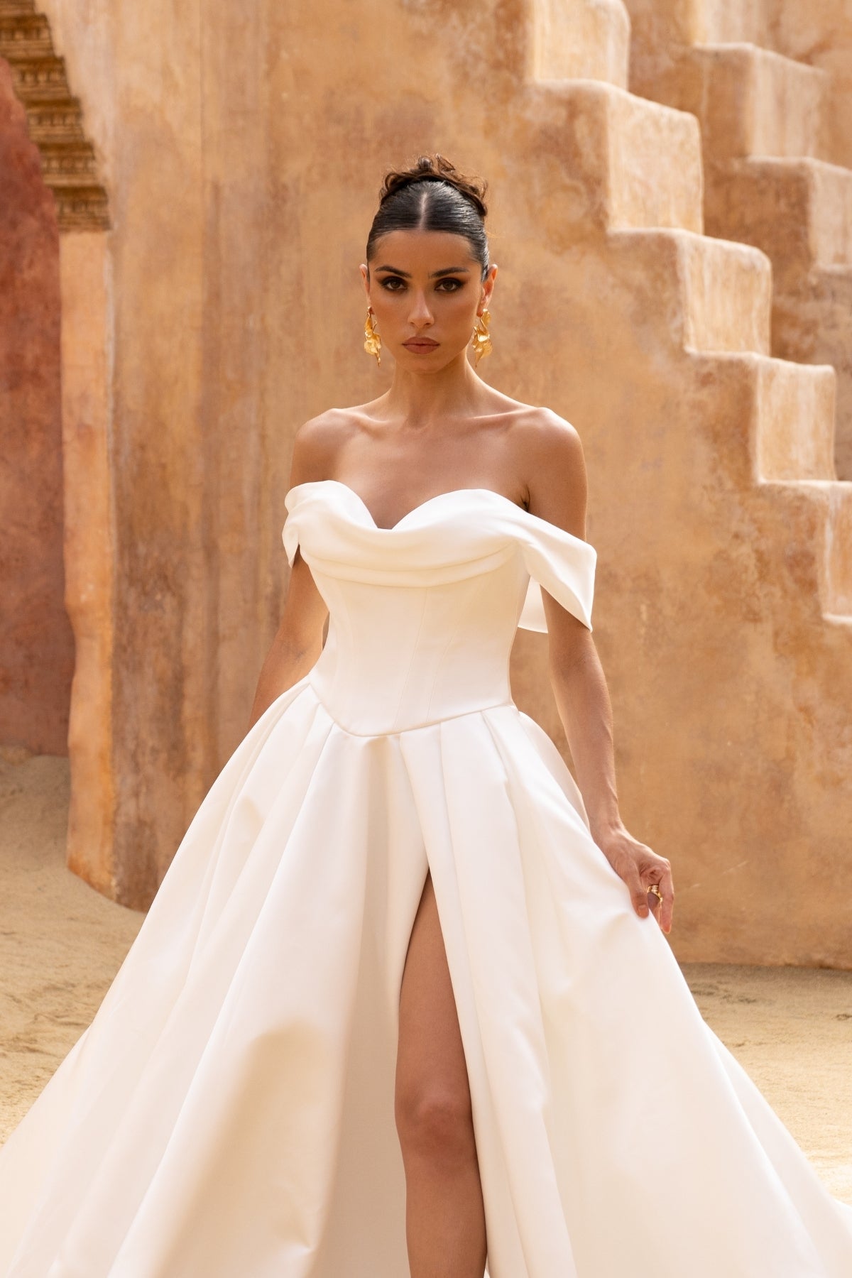 A line wedding gown featuring off the shoulder straps and a sweetheart neckline, in a minmal plain fabric, high split and dramatic skirt. 