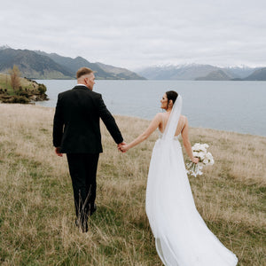 Bride and groom hold hands near lake, three metre veil trails behind.
