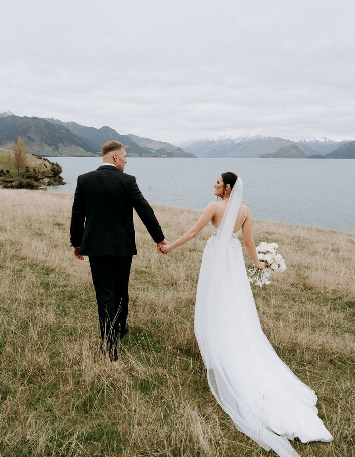 Bride and groom hold hands near lake, three metre veil trails behind.
