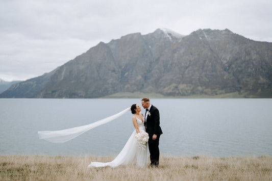Bride and groom stand in front of lake and mountain, veil flows in wind