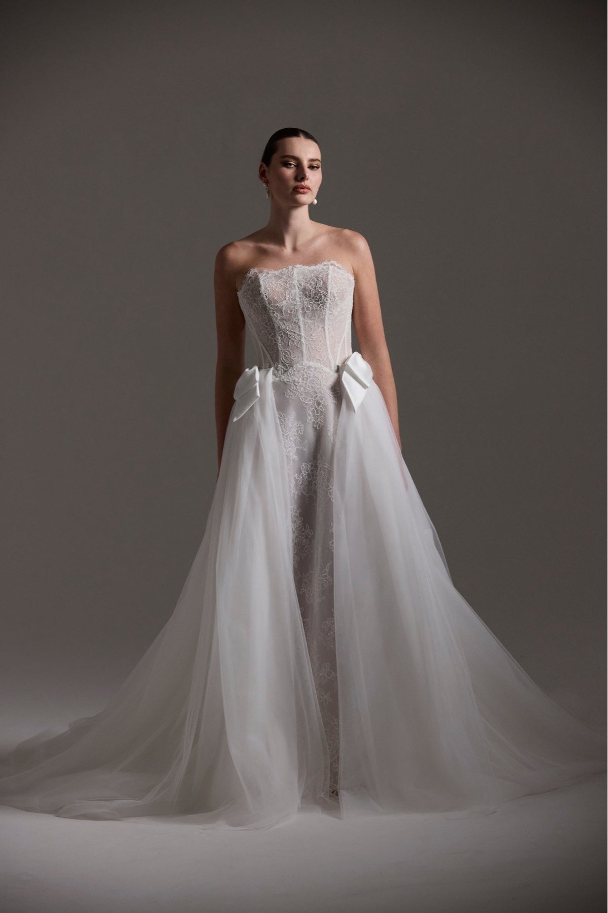 lace A-line wedding dress featuring a strapless neckline, exposed corset boning and unique waistline. worn with an Tulle overskirt adorned with ribbons on either side of hips. 