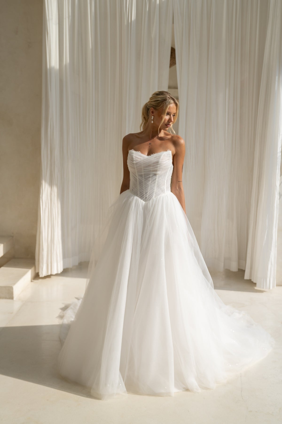 A-line wedding gown with cat-eye strapless neckline, criss cross ruching around the bust, corset boning on the bodice, basque waist and a voluminous tulle skirt. 