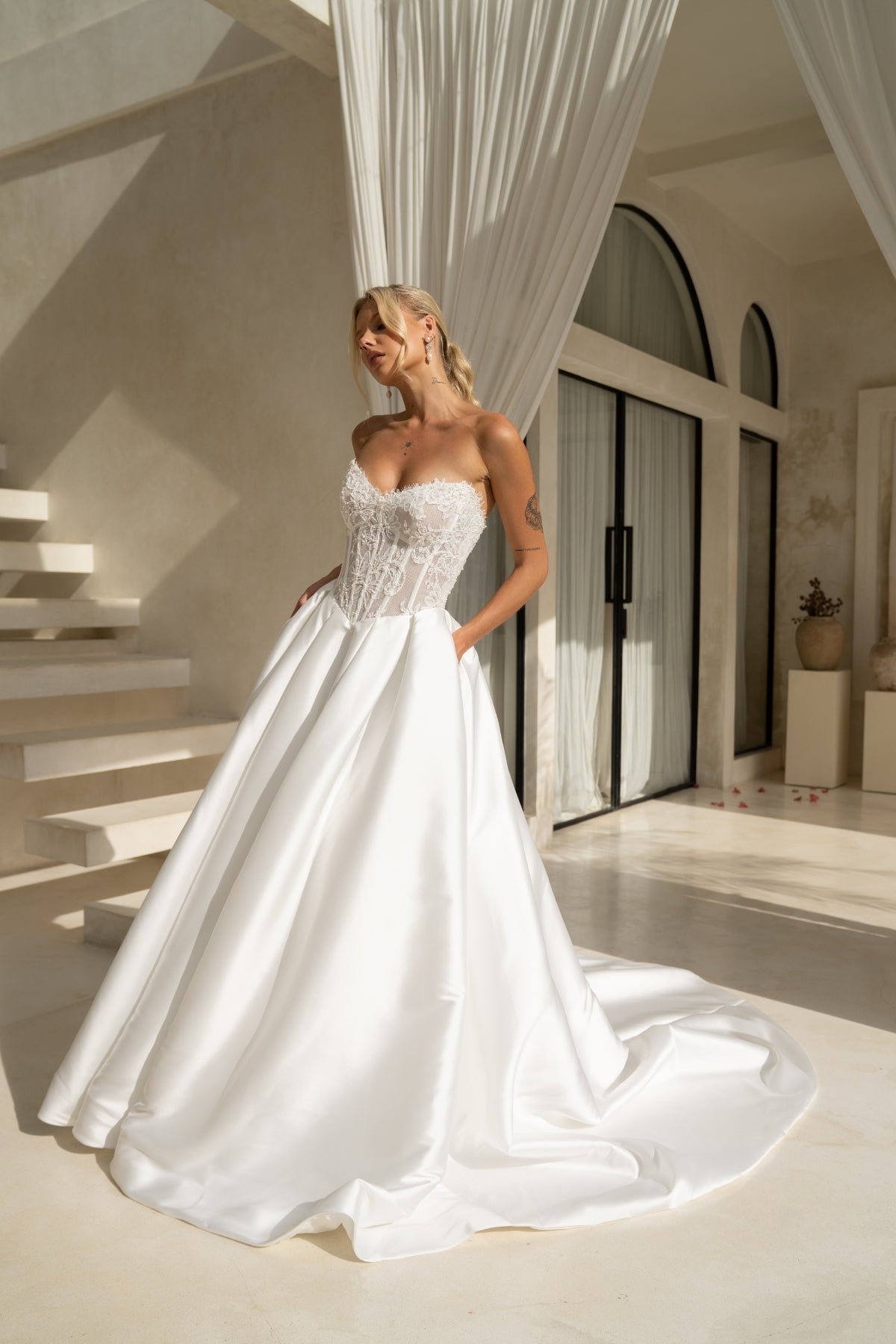 A-line wedding dress with a strapless sweetheart bodice covered in lace and corset boning, basque waistline, and a voluminous dramatic skirt in plain fabric. 