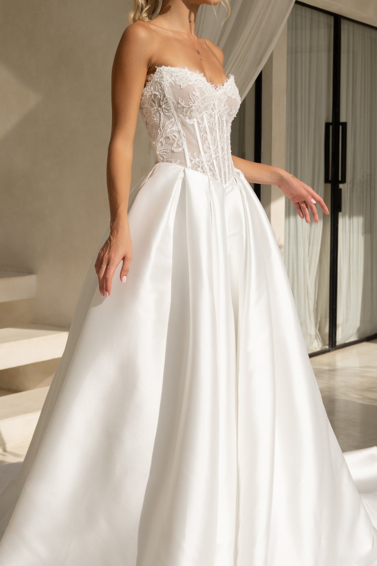 A-line wedding dress with a strapless sweetheart bodice covered in lace and corset boning, basque waistline, and a voluminous dramatic skirt in plain fabric. 