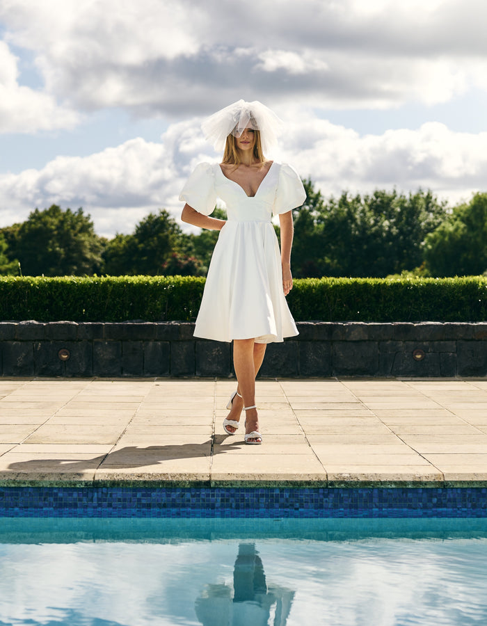 Bride stands in front of pool in mini dress and short fascinator-like veil