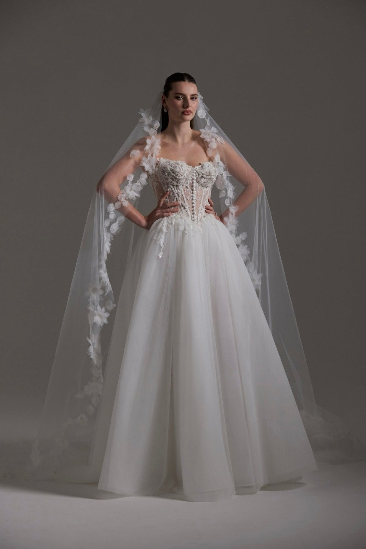 A-line Wedding dress featuring a tulle skirt, lace corseted bodice with basque waistline, cat-eye neckline, and buttons down the front of bodice. 
