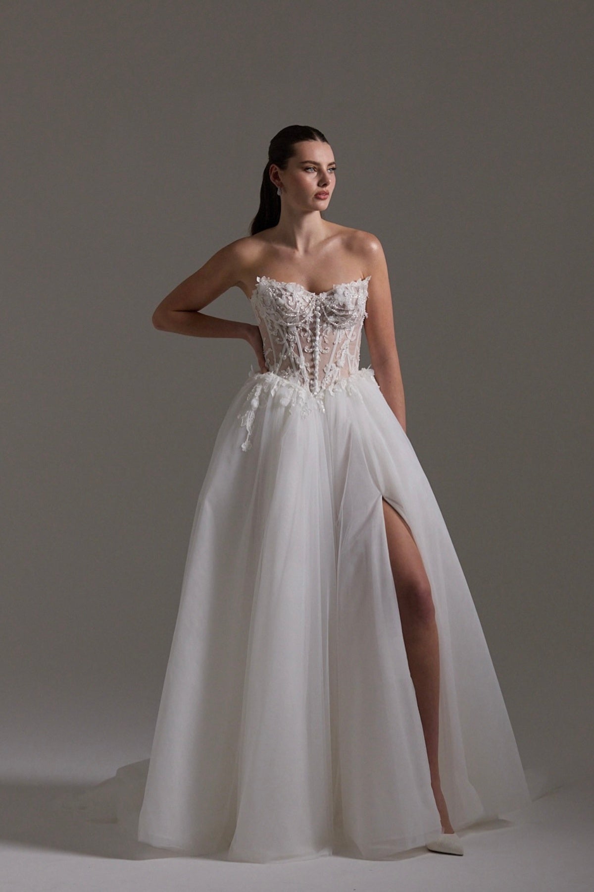 A-line Wedding dress featuring a tulle skirt, lace corseted bodice with basque waistline, cat-eye neckline, and buttons down the front of bodice. 