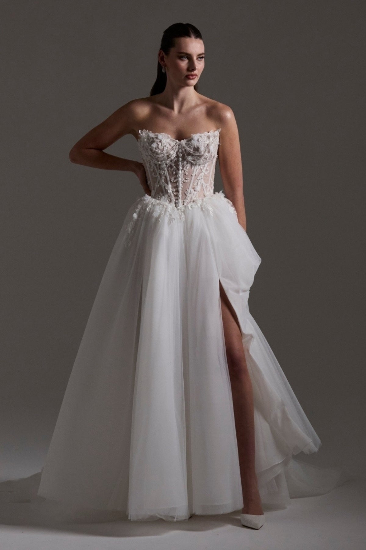 A-line Wedding dress featuring a tulle skirt, lace corseted bodice with basque waistline, cat-eye neckline, and buttons down the front of bodice. 