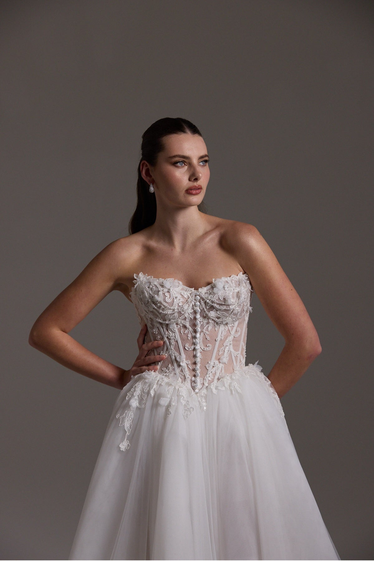 A-line Wedding dress featuring a tulle skirt, lace corseted bodice with basque waistline, cat-eye neckline, and buttons down the front of bodice. 