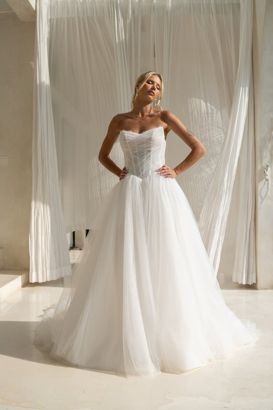 A-line wedding gown with cat-eye strapless neckline, criss cross ruching around the bust, corset boning on the bodice, basque waist and a voluminous tulle skirt. 