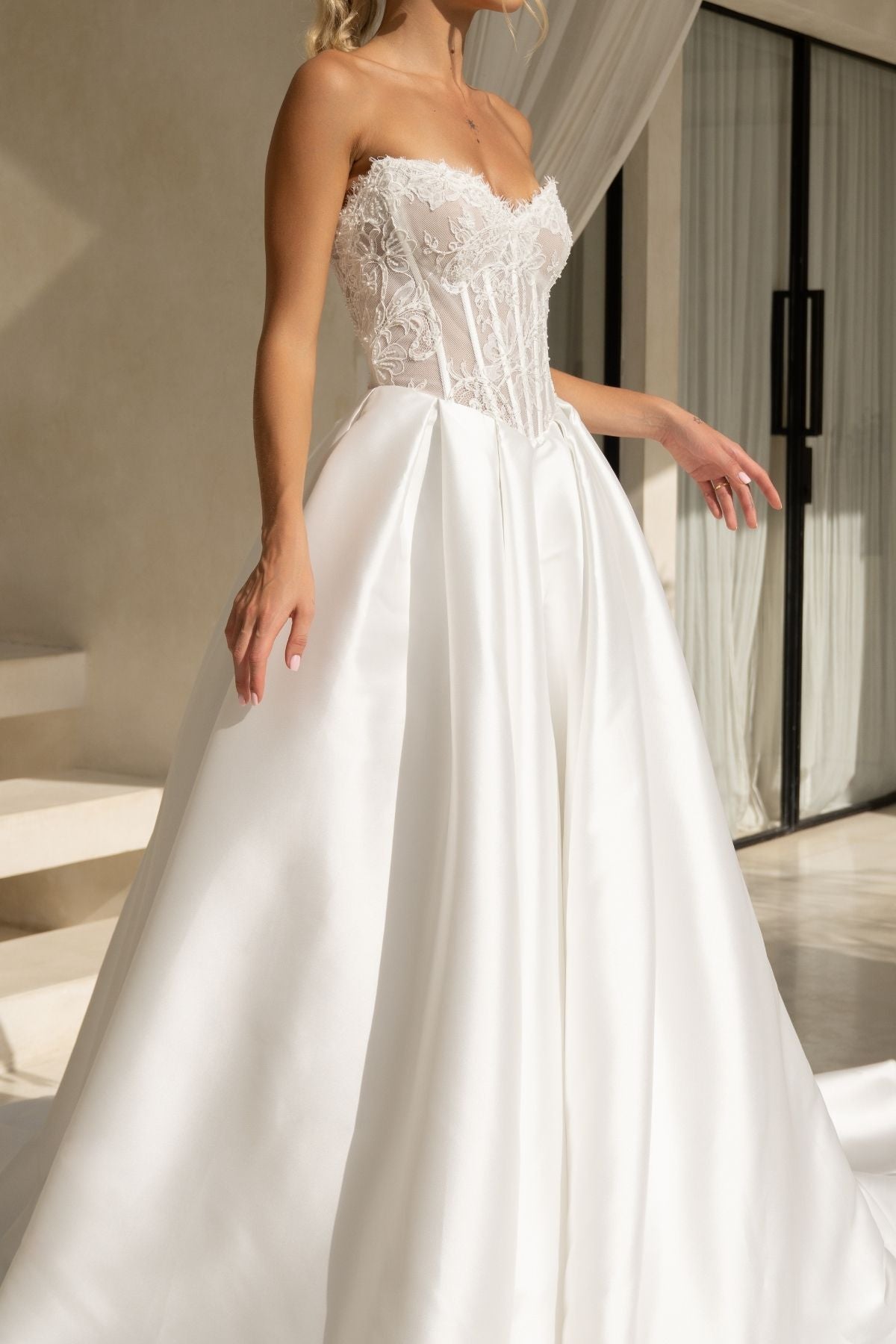 A-line wedding dress with a strapless sweetheart bodice covered in lace and corset boning, basque waistline, and a voluminous dramatic skirt in plain fabric. 