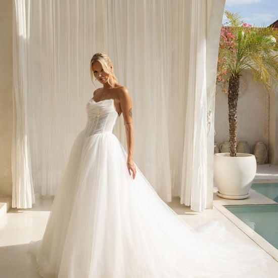 A-line wedding gown with cat-eye strapless neckline, criss cross ruching around the bust, corset boning on the bodice, basque waist and a voluminous tulle skirt. 