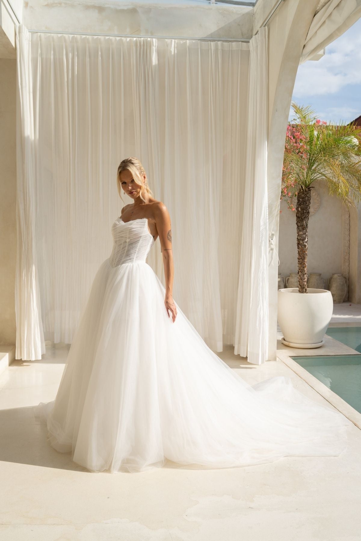 A-line wedding gown with cat-eye strapless neckline, criss cross ruching around the bust, corset boning on the bodice, basque waist and a voluminous tulle skirt. 
