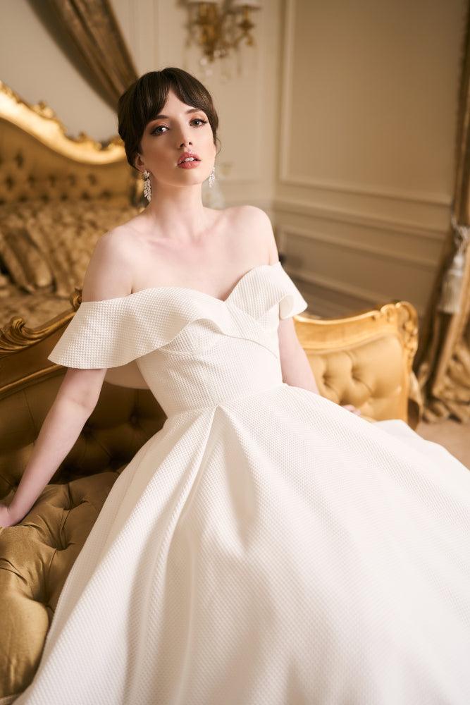 Tyler dress. Close up of front bodice. Model sits in voluminous ivory a-line dress made in a waffle jacquard fabric. Boned bodice with wavey off-shoulder band that crosses at front creating sweetheart neckline.