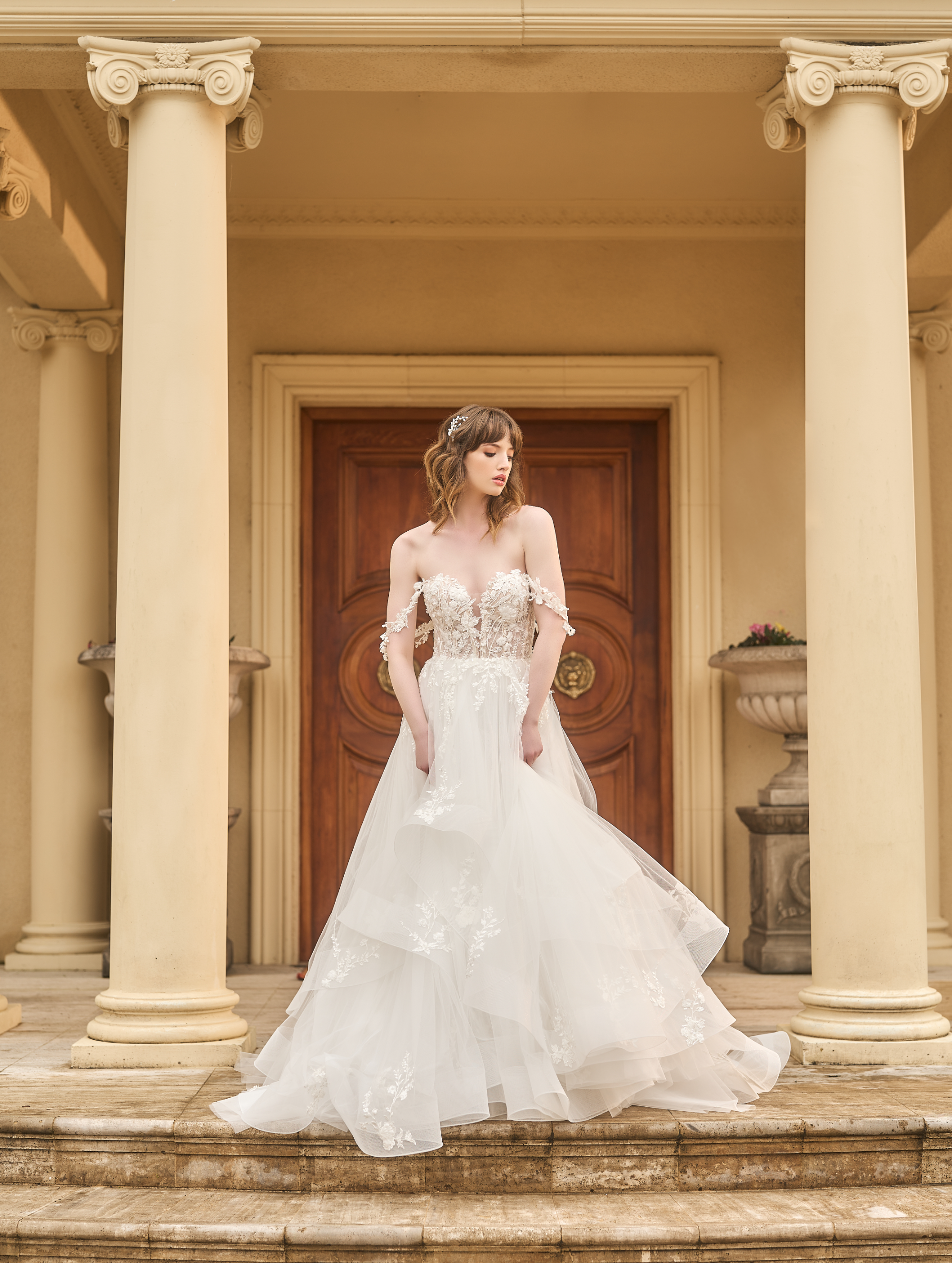 Tess is an a-line wedding gown with plunging sweetheart neckline. Asymmetrical tulle skirt creates volume. Delicate floral off-the-shoulder straps match the lace bodice and applique on skirt.