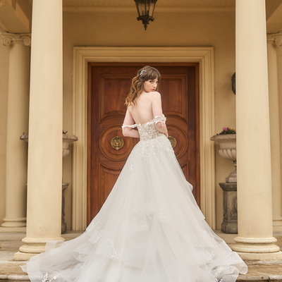 Back view of Tess. A-line wedding gown with boned bodice. Asymmetrical tulle skirt structured with horsehair hem. Delicate floral off-the-shoulder straps match the lace bodice and applique on skirt.