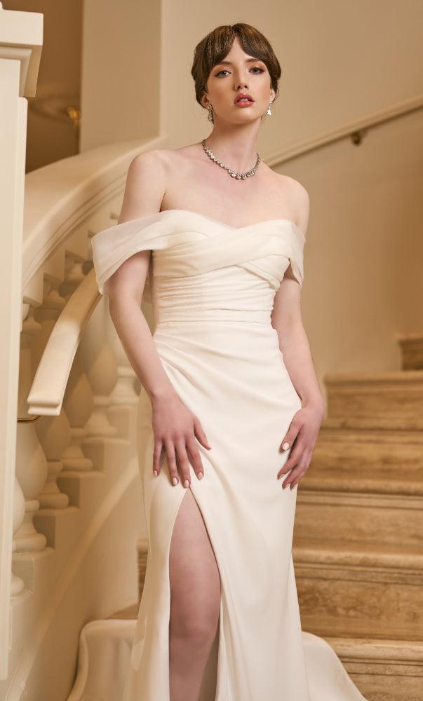 Close up of Tamsin bodice. Ivory crepe Tamsin wedding gown with ruched bodice and skirt with split over right leg. Organza crossover off-shoulder band creates sweetheart neckline.