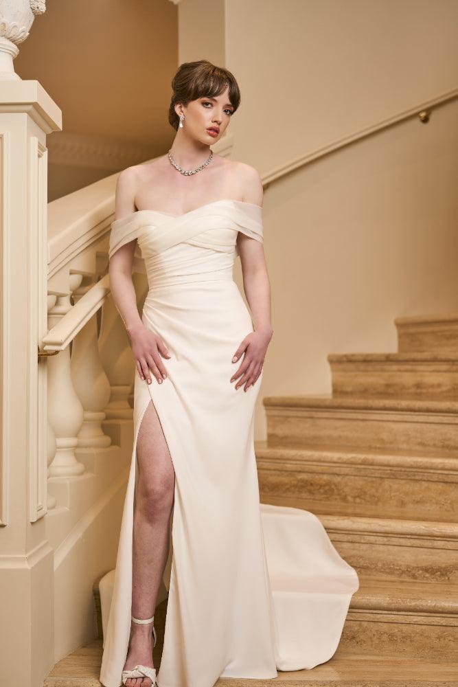 Ivory crepe Tamsin wedding gown with ruched bodice and skirt with split over right leg. Organza crossover off-shoulder band creates sweetheart neckline. Fit-n-flare silhouette with modest train.