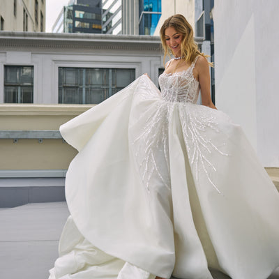 Raffael wedding dress from Jessica Coutures. Square neckline, low back A-line dress with organza fabric and glitter detailing at the bodice. Boning support, cinched waist, dramatic skirt. Hidden features include pockets and a split for easy movement.