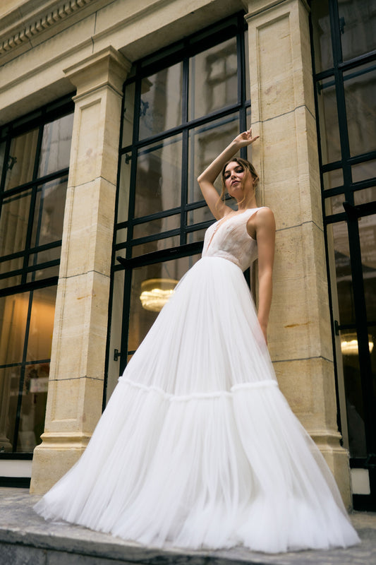 Rhia wedding dress with sheer pleated French Tulle fabric and A-line silhouette. Plunging v-neckline with floral lace overlay for a romantic touch. Two-tiered skirt adds volume and movement. Feminine and modern gown that gives the illusion of floating when walking down the aisle.