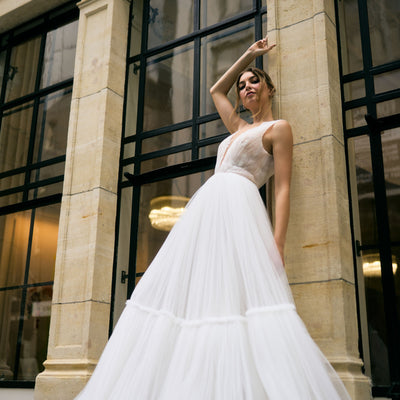 Rhia wedding dress with sheer pleated French Tulle fabric and A-line silhouette. Plunging v-neckline with floral lace overlay for a romantic touch. Two-tiered skirt adds volume and movement. Feminine and modern gown that gives the illusion of floating when walking down the aisle.