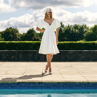 Model wears Poppi ivory minidress standing next to pool. Dress has A-line skirt and short balloon sleeves