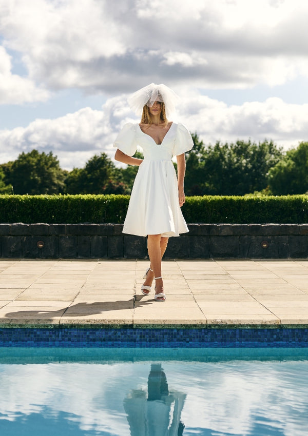 Model wears Poppi ivory minidress standing next to pool. Dress has A-line skirt and short balloon sleeves