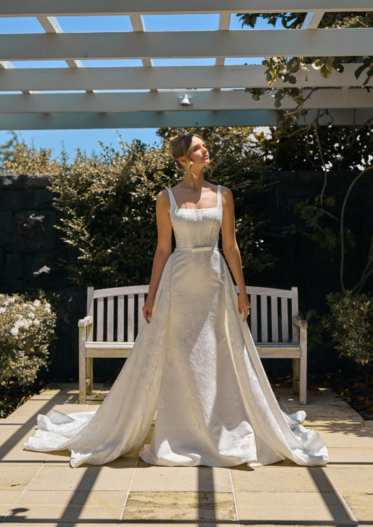 Model wears Perry dress with detachable perry overskirt in ivory jacquard fabric. 